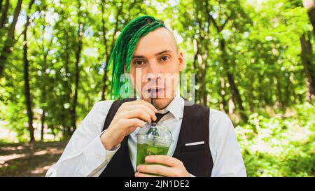 Junger Mann mit Dreadlocks trinkt erfrischende Limonade im Waldpark. Stilvoller Hipster im Business-Anzug mit grüner Frisur und alkoholfreien Getränken Stockfoto