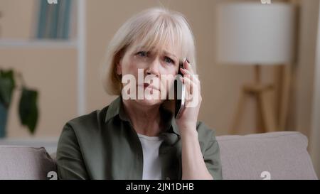Kaukasische alte Frau reifen mittleren Alters 50s beleidigte Dame im Alter Geschäftsfrau sitzen auf dem Sofa zu Hause hören Stimme auf dem Telefon sprechen Smartphone Stockfoto