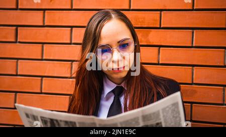 Junge Frau liest Zeitung auf der Straße. Dame mit langen Haaren in legerer Kleidung, die auf der Straße steht und Zeitung liest. Stockfoto