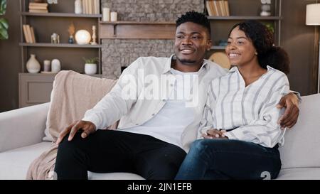 Glückliches, frisch verheiratetes Paar, afrikanischer Mann und Frau, die auf dem Sofa zu Hause sitzen und Fernsehen und lachen. Ehemann und Ehefrau sprechen mit Familientherapeuten Stockfoto