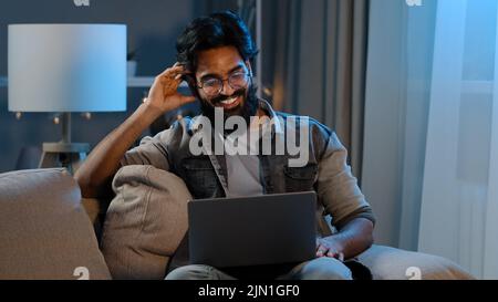 Arabisch lächelnd glücklich bärtig Mann Millennial Kerl Geschäftsmann sitzt auf dem Sofa zu Hause in der Nacht dunkel spät winkt hallo in webcam Online-Video Stockfoto