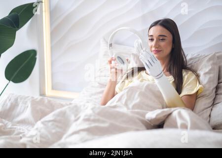 Glückliches Mädchen mit bionischen Arm entspannend Musik hören Kopfhörer im Bett zu Hause. Stockfoto