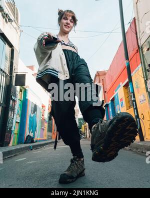 Trendige Transgender kaukasische junge lateinische Frau trägt eine Jacke und baggy Hosen, auf der Straße stehend posiert heben ein Bein, vertikales Bild. Stockfoto