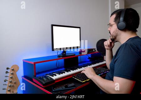 kaukasischer latein mit Kopfhörern und Brille, der in seinem Heimmusikstudio sitzt und mit der Hand auf dem Kinn denkt und auf den Monitor schaut, Mus Stockfoto