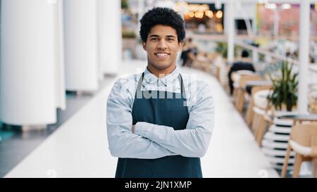 Junger afroamerikanischer Mann Kerl Restaurant Café männlich Arbeiter Blick auf Kamera posiert Kreuzung Arme Kellner Verkäufer trägt Schürze stehen am Arbeitsplatz Stockfoto