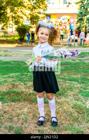 Lysytschansk, Ukraine. 1. September 2021 - ein Mädchen mit einem Blumenstrauß in einer weißen Bluse mit Schleifen auf dem Hintergrund der Schule während der Stockfoto