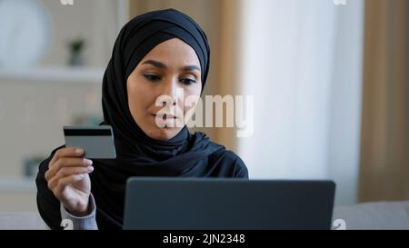 Lächelndes islamisches Mädchen Kunde in Hijab sitzt auf dem Sofa Online-Bestellung verwenden Laptop geben Kreditkartennummer für Transaktion glücklich muslimische Frau zahlen Stockfoto