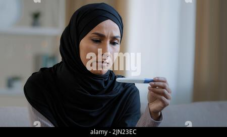 Schockiert unglücklich islamische junge Frau arabisch muslimische Mädchen in Hijab weiblichen Erwachsenen schwanger Gefühl Angst Blick auf positive Schwangerschaftstest Ergebnis sitzen an Stockfoto