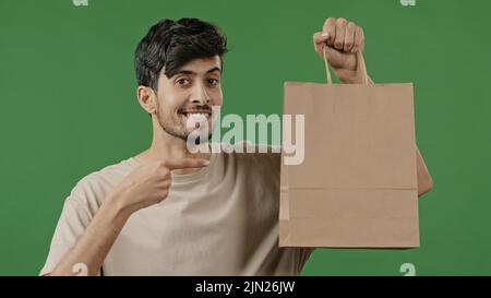 Portrait junger lächelnder Kerl mit Papierpaket, der Finger auf Einkaufstasche zeigt hispanic-männlicher Käufer, der im Studio auf grünem Hintergrund steht, freut sich Stockfoto