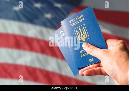 Zwei biometrische ukrainische Pässe in der Hand eines Mannes vor dem Hintergrund der Nationalflagge der Vereinigten Staaten von Amerika. Reisen, Visa, Emigrati Stockfoto