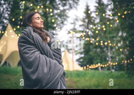 Eine junge Frau, die in eine Decke im Wald auf einem verschwommenen Hintergrund gehüllt ist. Stockfoto