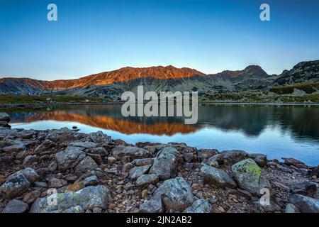 Sonnenaufgang mit sanftem, orangefarbenem Licht, das die Gipfel des Retezat-Gebirges erhellt und sich im Bucura-See widerspiegelt. Foto aufgenommen am 7.. August 2022 am Ufer Stockfoto