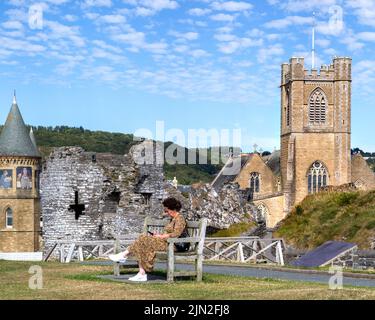 Eine Frau schaut auf ihr Telefon, während sie auf einer Bank auf dem Gelände des Schlosses Aberystwyth sitzt. Die St. Michaelskirche ist über einen Teil der Burgruine hinaus zu sehen. Stockfoto