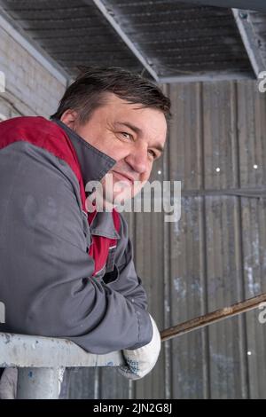 Ein müder Arbeiter mittleren Alters in einer schmutzigen Schutzuniform schaut in die Ferne, während er in einem Lagerhaus, einer Fabrik und einem vertikalen Rahmen arbeitet. Hochformat Stockfoto