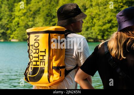 RIJEKA, KROATIEN - 10 2022. JULI: Touristen mit dunklen Panamas wandern entlang des blauen Sees, umgeben von Bäumen. Mann mit gelbem Rucksack The North Face und Frau auf Exkursion Blick auf den See zurück Stockfoto