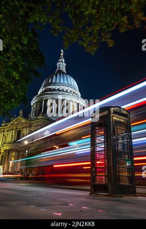 London, Großbritannien - 11. Juni 2022: Leichte Wege eines roten Doppeldeckerbusses hinter einer schwarzen Telefonzelle mit der Kuppel der St. Paul's Cathedral im Hintergrund Stockfoto