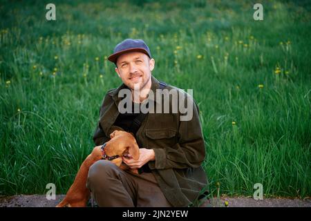 Netter Rüde, der im Park mit grünem Gras sitzt und mit einem kleinen braunen Welpen spielt. Haustiere als Familienmitglieder Konzept Bild. Schöner junger Mann mit Dackel im Freien. Mann auf grünem Gras mit Hund Stockfoto