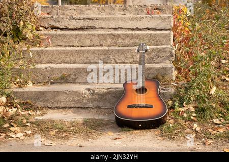 Alte braune Gitarre liegt im Park auf den Stufen im Herbst in der Sonne, Gitarre Stockfoto