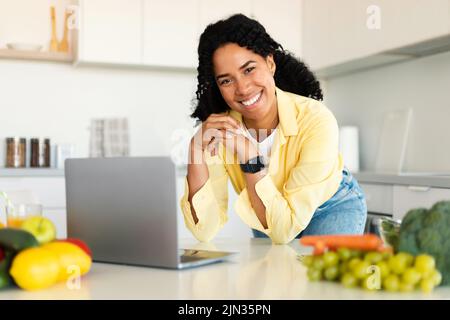 Neue Rezepte werden gesucht. Glückliche afroamerikanische Frau, die in der Küche einen Laptop benutzt, während sie frischen Salat kocht Stockfoto