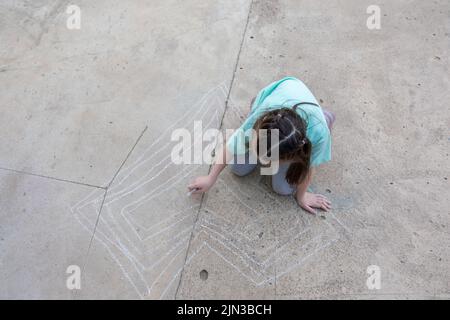 Mädchen zeichnet mit bunten Buntstiften auf dem Bürgersteig. Kinderzeichnungen mit Kreide an der Wand. Kreatives Kind. Freude an der Kindheit. Hochwertige Fotos Stockfoto