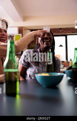 Fröhliche multiethnische Frauen spielen Karte raten Spiel mit Freunden im Wohnzimmer. Eine Gruppe verschiedener Freunde, die im Wohnzimmer am Tisch sitzen und gemeinsam Gesellschaftsspiele spielen. Stockfoto