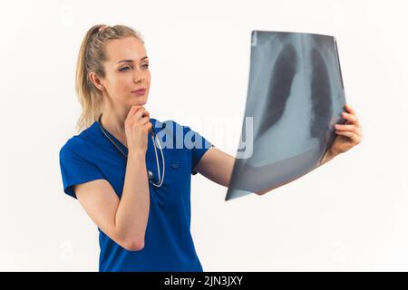 Kaukasische blonde Ärztin mit Pferdeschwanz und Stethoskop, die Röntgenaufnahmen der Lunge untersucht und dabei die Hand auf das Kinn fokussiert sieht. Studioaufnahme. Isolierter Kopierbereich. Hochwertige Fotos Stockfoto