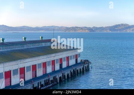 Lagerhaus am Pier in Fisherman's Wharf in San Francisco, Kalifornien. Altes Lagerhaus mit roten Toren gegen den Blick auf die Bucht und die Berge. Stockfoto