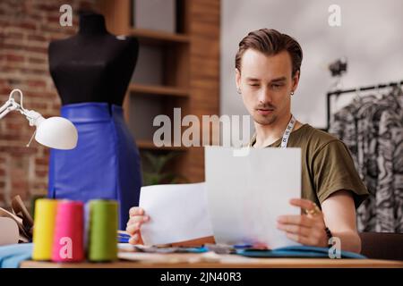 Ein junger Mann ist ein Modedesigner. Er schaut sich die Skizze an, während er mit einer Nähmaschine an einem Tisch sitzt Stockfoto