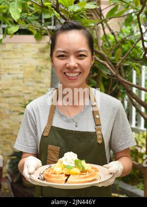Die vietnamesische Kellnerin serviert belgische Waffeln mit Obst und Eis im Café im Freien Stockfoto