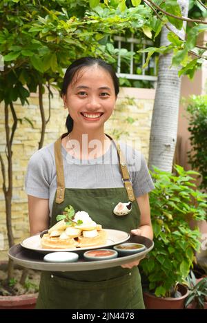 Die vietnamesische Kellnerin serviert belgische Waffeln mit Obst und Eis im Café im Freien Stockfoto