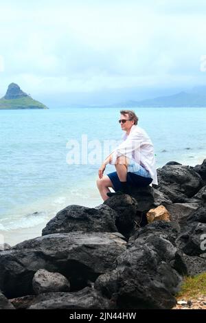 Attraktiver weißer Mann aus den Vierzigern, der am felsigen Ufer des hawaiianischen Strands bei Mokoli'i Island (früher bekannt als der veraltete Begriff „Chinaman's hat“) sitzt Stockfoto