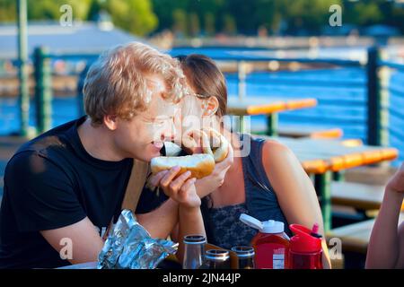 Junges Paar, das Hotdogs am See isst und Spaß hat Stockfoto