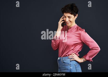 Hallo wie kann ich helfen. Eine attraktive junge Geschäftsfrau steht und spricht auf ihrem Handy vor schwarzem Hintergrund im Studio. Stockfoto