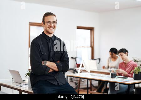 Die Zukunft lässt sich durch die Schaffung vorhersagen. Ein beschnittenes Porträt eines hübschen jungen Geschäftsmannes, der mit gefalteten Armen sitzt, während seine Kollegen sitzen Stockfoto