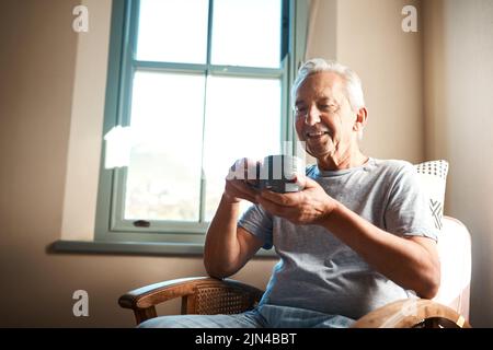 Hmm, nichts wie eine Tasse Kaffee am Morgen. Ein glücklicher älterer Mann hält eine Tasse Kaffee und sitzt in einem Sessel zu Hause. Stockfoto