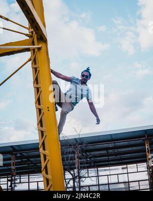 Lateinischer junger Mann, der Parkour auf einer gelben Metallstruktur übt. Hispanischer Junge in einem blauen Hemd, der auf einer gelben Metallstruktur klettert. Junger Mann c Stockfoto