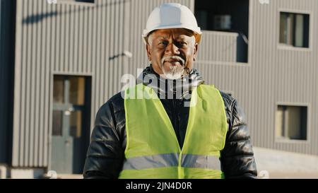 Nahaufnahme älterer afroamerikanischer Bauarbeiter, der in Schutzhelm-Uniform im Freien stand, selbstbewusster Arbeiter kreuzte die Arme über die Brust Stockfoto