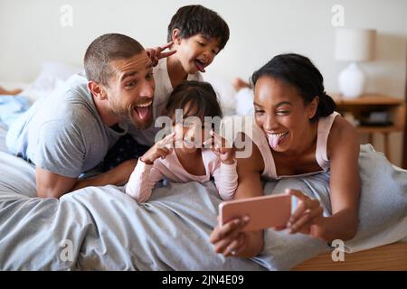 Alberne Gesichter und Selfies am Morgen. Eine wunderschöne junge vierköpfige Familie, die mit einem Handy zu Hause in ihrem Schlafzimmer ein Selfie macht. Stockfoto