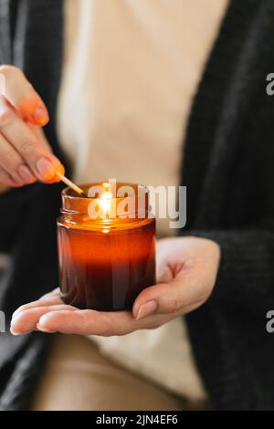 Frau hält die Kerze in der Hand und zündet die Kerze in einem kleinen Glas aus Bernsteinglas mit Holzdocht an. Zen- und Relax-Konzept. Stockfoto