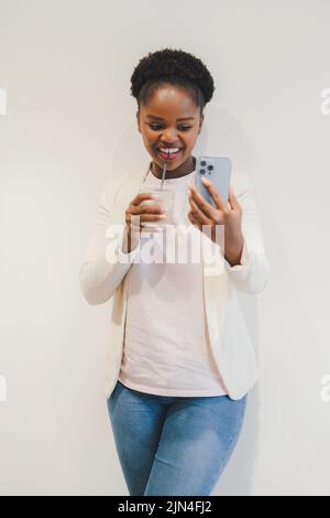 Lächelnde afro-amerikanische Frau, die einen Videoanruf auf dem Handy macht, ein Glas Getränk trinkt, isoliert auf weißem Hintergrund. Kreative neue Idee und Stockfoto