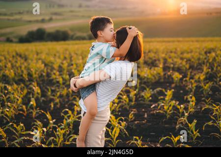 Mutter hält das Kind in den Armen, während er sie auf die Stirn küsst. Fotografieren im Maisfeld. Unbeschwerte Kindheit. Stockfoto