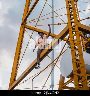 Junger hispanischer Junge in einem gelben Hemd, der hoch oben auf einer gelben Struktur sitzt. Gelber Industrieturm mit einem Jungen beim Klettern. Junge oben mit den s Stockfoto