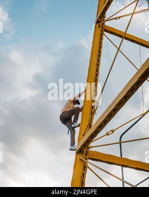 Athletischer hispanischer Mann mit dem Himmel im Hintergrund, der eine Metallstruktur klettert. Ein junger Mann in Stadtkleidung und einem gelben Hemd, der zum Aufstieg Stockfoto