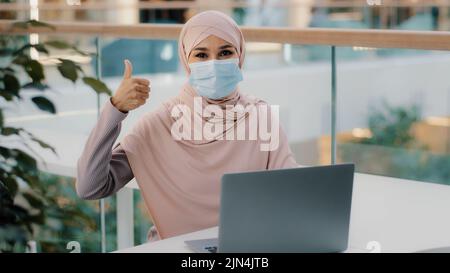 Glückliche junge arabische Geschäftsfrau, die am Schreibtisch sitzt und auf einem Laptop am Internet tippt und lächelnd die neue App überprüft und dabei den Daumen der Kamera zeigt Stockfoto