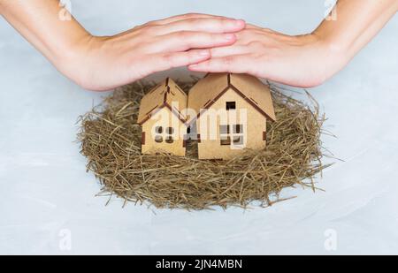 Die Hände bedecken zwei Miniatur-Holzhäuser, die in einem auf neutralem Hintergrund isolierten Heuennest platziert sind. Hausversicherung und Schutzkonzept. Stockfoto