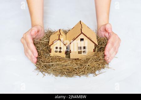 Weibliche Hände, die ein Heuennest mit großen und kleinen Häusern auf neutralem Hintergrund machen. Sicheres Familienhauskonzept. Stockfoto