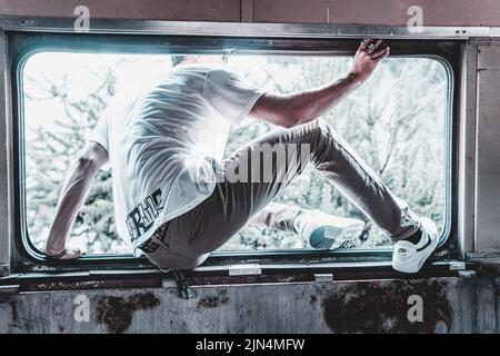 In Weiß gekleideter Junge, der aus dem Fenster in einem verlassenen Gebäude klettert. Hispanischer Mann, der einen verlassenen Ort erkundet. Lateinischer junger Junge, der durch die kommt Stockfoto