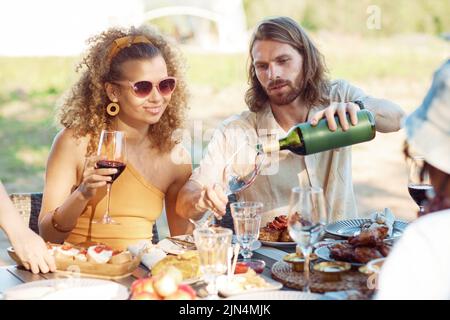 Porträt eines jungen Paares, das im Sommer Rotwein ins Glas gießt, während es im Freien eine Dinnerparty mit Freunden genießt Stockfoto