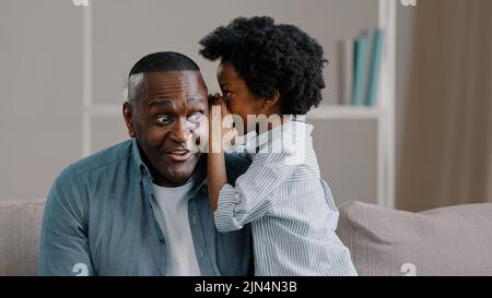 Kleine süße Tochter flüstert in Dad's Ohr erzählen Geheimnis sagt lustig Witz liebevollen Vater mit Kind Mädchen Spaß auf der Couch im Zimmer sitzen Stockfoto