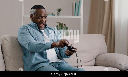 Aufgeregt besorgt emotionale Erwachsene afroamerikanische männliche Gamer auf dem Sofa im Wohnzimmer sitzend Videospiel genießt Hobby halten Controller Joystick spielen Stockfoto